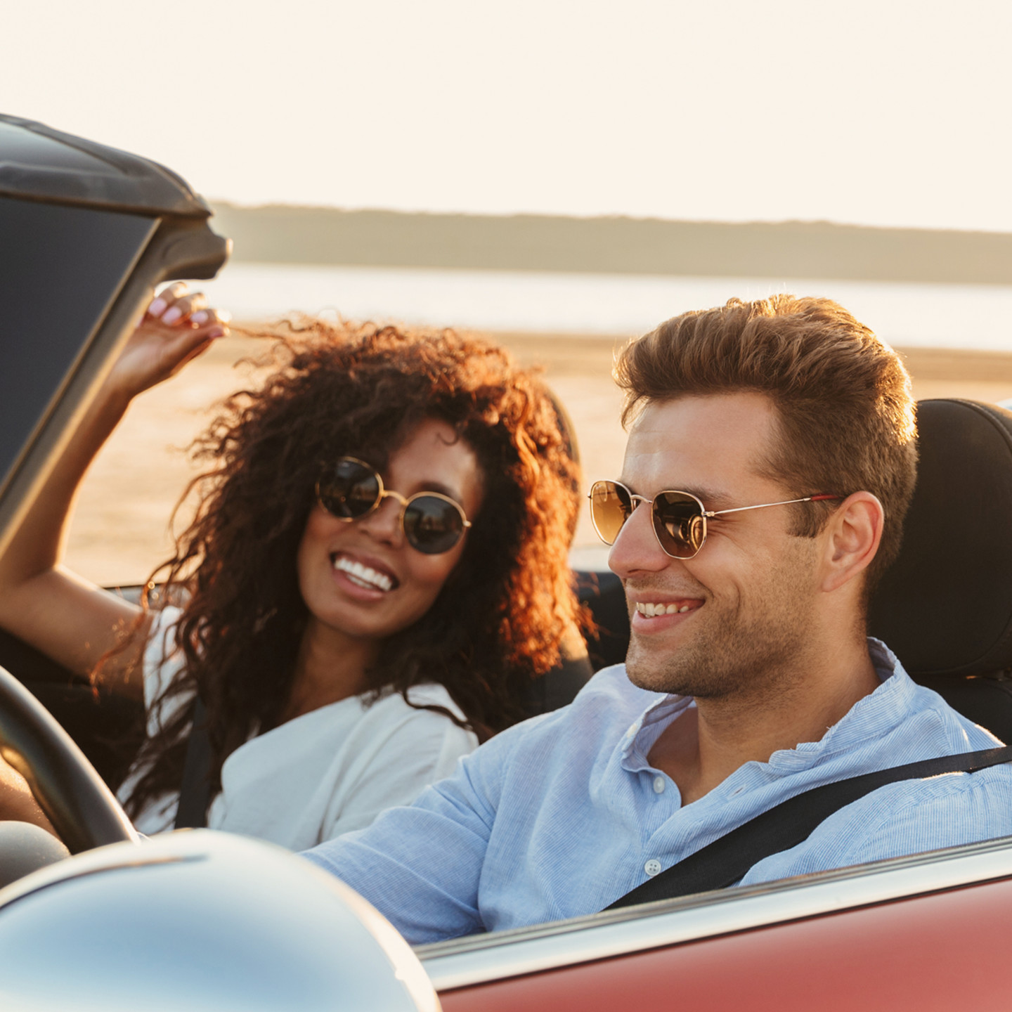 Mann und Frau im Cabrio, beide mit runder Sonnenbrille, im Hintergrund Strand und Meer.